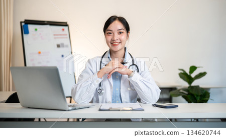 Asian female doctor holding pen with both hands under chin with a laptop on table in diagnosis room. 134620744