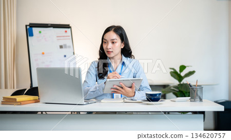 Female office worker businesswoman holding stylus writing in tablet while looking at laptop on table Female office worker businesswoman holding stylus writing in tablet while looking at laptop on table 134620757