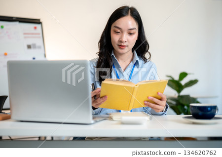 Female office worker or businesswoman reading yellow cover book with laptop aside on working table. Female office worker or businesswoman reading yellow cover book with laptop aside on working table. 134620762