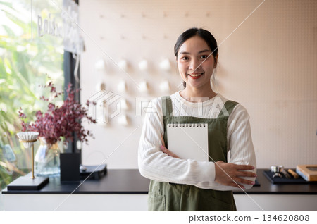 Asian female barista in green apron holding or hugging notebook waiting for customer at counter bar. Asian female barista in green apron holding or hugging notebook waiting for customer at counter bar. 134620808