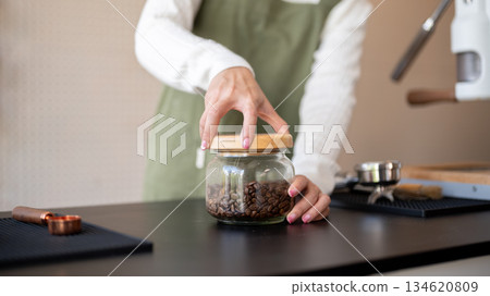 Close up of green apron barista hand opening or closing lid of coffee beans jar at cafe counter bar. Close up of green apron barista hand opening or closing lid of coffee beans jar at cafe counter bar. 134620809