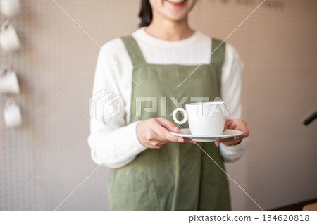 Asian female barista in green apron holding a cup of coffee serving to customer at cafe counter bar. 134620818