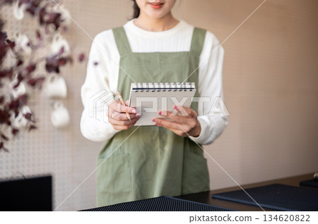 Asian female barista in green apron holding pencil and notebook checking order at cafe counter bar. Asian female barista in green apron holding pencil and notebook checking order at cafe counter bar. 134620822