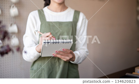 Asian female barista writing in notebook with pencil taking a customer's order at cafe counter bar. Asian female barista writing in notebook with pencil taking a customer's order at cafe counter bar. 134620823