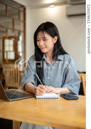 Smiling asian woman holding pen writing in notebook while looking at laptop on cafe's wooden table. Smiling asian woman holding pen writing in notebook while looking at laptop on cafe's wooden table. 134620972