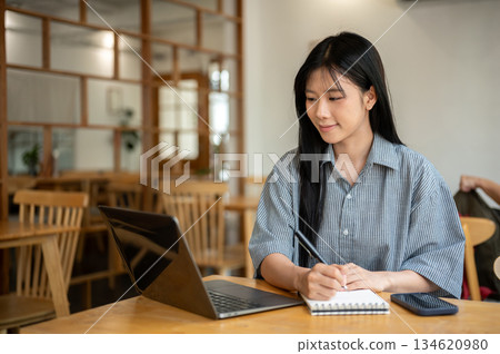 Smiling asian woman holding pen writing in notebook while looking at laptop on cafe's wooden table. 134620980