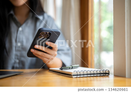Close up of woman's hand holding or touching on phone with notebook and laptop at cafe wooden table. 134620981