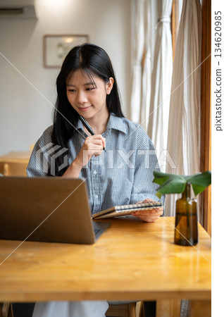 Asian woman holding notebook and pen while looking at laptop with vase aside on cafe's wooden table. Asian woman holding notebook and pen while looking at laptop with vase aside on cafe's wooden table. 134620985