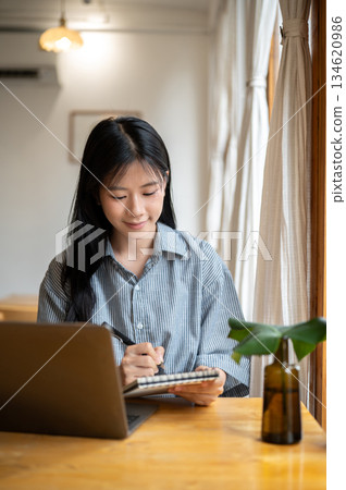 Smiling asian woman holding a pen and writing in notebook with laptop aside on cafe's wooden table. Smiling asian woman holding a pen and writing in notebook with laptop aside on cafe's wooden table. 134620986