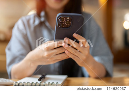 Close up of woman hold phone in both hands watching video or texting message over a notebook on cafe 134620987
