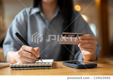 Close up of a woman holding credit or debit card and a pen writing in notebook sitting at cafe table Close up of a woman holding credit or debit card and a pen writing in notebook sitting at cafe table 134620988