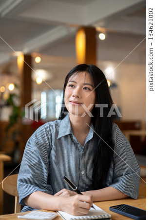 Asian woman looking away as holding pen over notebook with calculator and phone on cafe wooden table 134621002