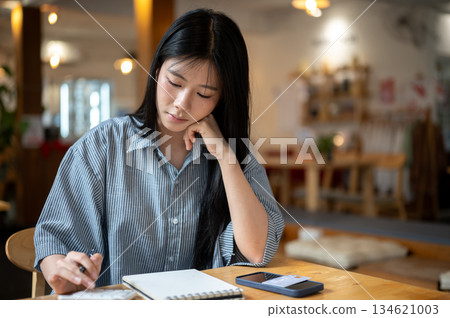 Asian woman putting hand under chin looking at calculator with notebook aside on cafe's wooden table Asian woman putting hand under chin looking at calculator with notebook aside on cafe's wooden table 134621003