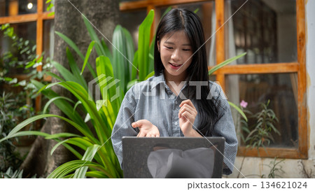 Smiling asian woman explaining with her hands while looking at laptop and sitting at cafe's outdoor garden. Smiling asian woman explaining with her hands while looking at laptop and sitting at cafe's outdoor garden. 134621024
