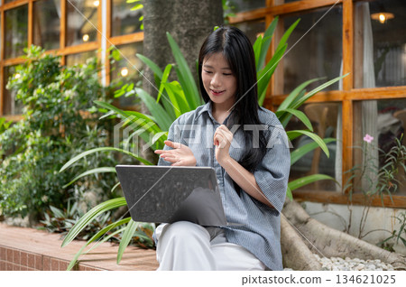 Asian woman explaining with hand while sitting cross legged looking at laptop at cafe outdoor garden Asian woman explaining with hand while sitting cross legged looking at laptop at cafe outdoor garden 134621025