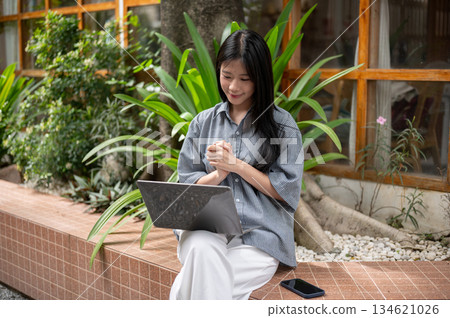 Smiling asian woman clasping her hands while sitting cross legged and looking at laptop at cafe's outdoor garden 134621026