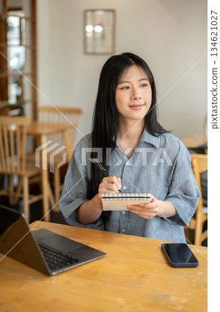 Smiling asian woman holding pen looking away from notebook with laptop aside on wooden table in cafe Smiling asian woman holding pen looking away from notebook with laptop aside on wooden table in cafe 134621027