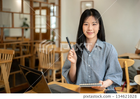 Smiling asian woman holding pen up looking away from notebook with laptop aside on cafe wooden table 134621028