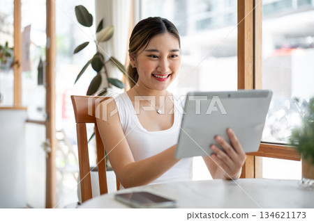 Asian woman holding and looking at the tablet while sitting at round table beside glass wall in cafe Asian woman holding and looking at the tablet while sitting at round table beside glass wall in cafe 134621173