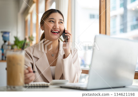 Asian woman talking on phone with pen over notebook sitting with laptop and coffee at cafe's table 134621198