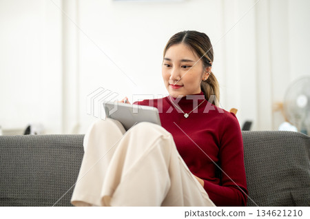 Asian woman designer holds stylus looking at tablet sitting legs up on sofa in living room or studio 134621210