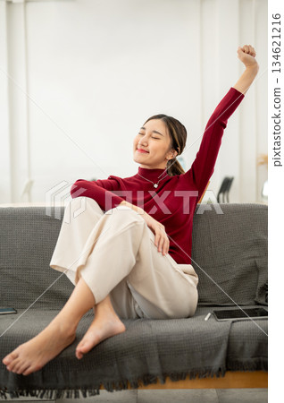 Pretty asian woman raising arm stretching as sitting legs up on fabric sofa in living room or studio 134621216