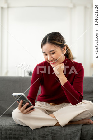 Pretty asian woman smiling as looking at phone fist under chin while sitting on sofa in living room. 134621224