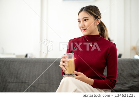 Asian woman smiling as holding ice coffee and sitting cross legged on sofa in living room or lounge 134621225