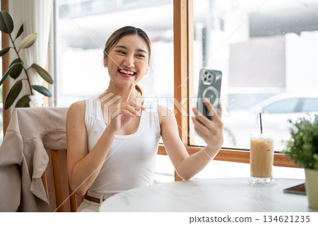 Asian woman holding phone talking on video call with finger pointing at cafe table with iced coffee. Asian woman holding phone talking on video call with finger pointing at cafe table with iced coffee. 134621235