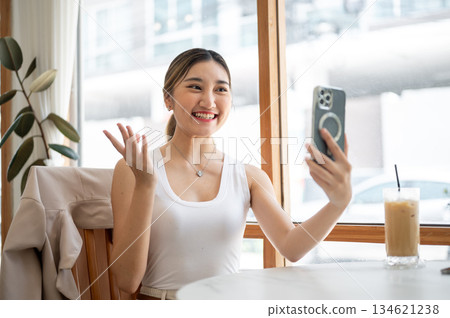Asian woman holding phone talking on video call with hand raising over cafe's table with iced coffee 134621238