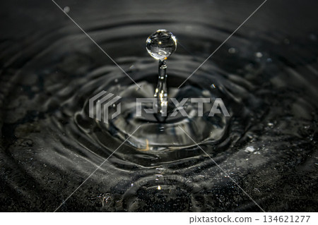macro capture of a water droplet rising from a narrow column, forming concentric ripples on a dark, reflective surface. Ideal for science, nature, and beautiful visuals macro capture of a water droplet rising from a narrow column, forming concentric ripples on a dark, reflective surface. Ideal for science, nature, and beautiful visuals 134621277