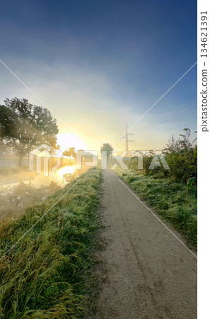 Enjoy a Serene Sunrise Walk Along a Misty Path by the Water, Where Nature Awaits Enjoy a Serene Sunrise Walk Along a Misty Path by the Water, Where Nature Awaits 134621391