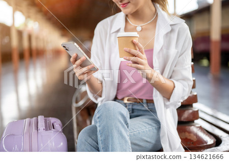 Close up of a woman holding coffee cup looking at phone as sitting on wooden bench at train station. 134621666
