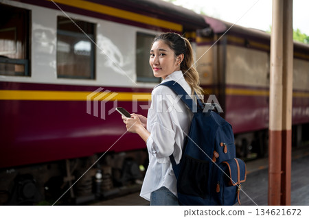 Asian woman with travel backpack holding phone looking back while standing waiting at train station. 134621672
