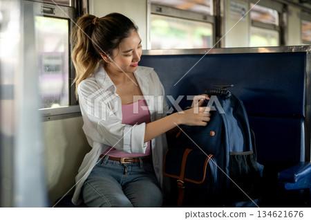 Pretty asian woman opening or zipping up blue bag while sitting on carriage bench at train station. 134621676