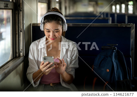 Asian woman wears headphone looking or choosing music as sitting on carriage bench at train station. 134621677