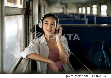 Asian woman wears headphone listening to music as sitting on bench and looking out at train station. 134621679
