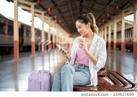 Pretty asian woman holding coffee as looking at phone while sitting on wooden bench at train station 134621680