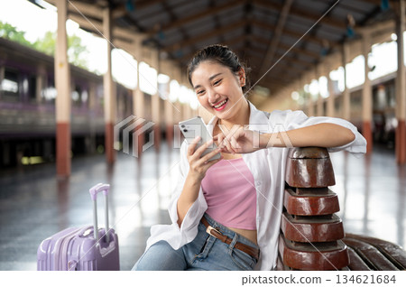 Pretty asian woman holding phone looking laughing as sit resting arm on train station's wooden bench Pretty asian woman holding phone looking laughing as sit resting arm on train station's wooden bench 134621684