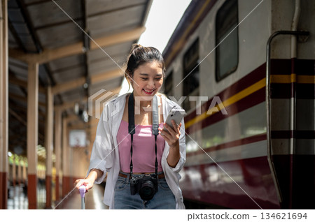 Asian woman smiling as looking at phone and holding luggage aside carriage at train station platform Asian woman smiling as looking at phone and holding luggage aside carriage at train station platform 134621694