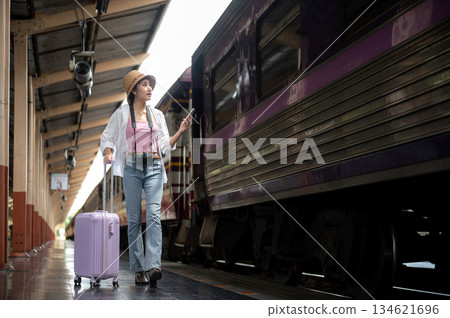 Asian woman holding phone looking at purple carriage and dragging luggage on train station platform. 134621696