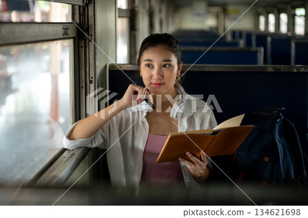 Pretty asian woman holding pen look away from book as sitting on bench in carriage at train station. Pretty asian woman holding pen look away from book as sitting on bench in carriage at train station. 134621698