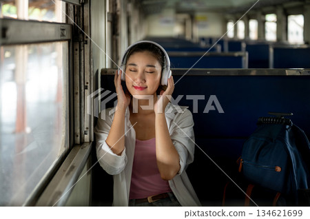Pretty asian woman wearing headphone listening to music sitting on carriage bench at train station. 134621699
