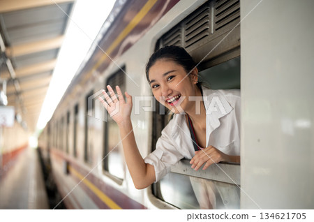 Smiling asian woman leaning head and waving hand out of carriage's window at train station platform Smiling asian woman leaning head and waving hand out of carriage's window at train station platform 134621705