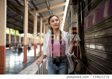 Pretty asian woman holding phone looking at carriage as dragging luggage on train station platform. Pretty asian woman holding phone looking at carriage as dragging luggage on train station platform. 134621710
