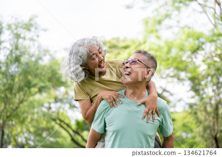 Glasses old man carrying old woman on his back and smiling at each other while standing in the park. 134621764