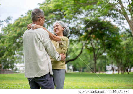 Old woman smiling and holding onto old man shoulders while dancing together on grass lawn in park. 134621771