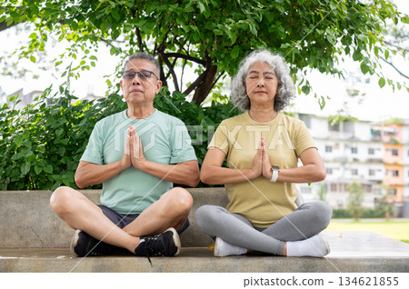 Glass old man and a woman sitting with joining palms and meditating together on cement bench in park Glass old man and a woman sitting with joining palms and meditating together on cement bench in park 134621855