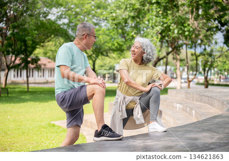 Old woman looking laughing at old man while stretching their legs and arms before exercise in park. Old woman looking laughing at old man while stretching their legs and arms before exercise in park. 134621863