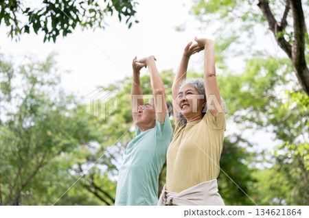Old man and woman smiling while standing and stretching their arms above head together in the park. 134621864